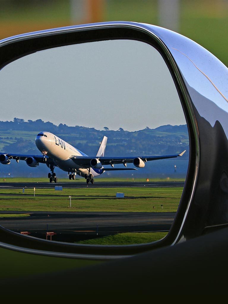 View through a car’s side mirror of a commercial airplane landing on a runway. The plane’s wheels are touching down, and the background shows a grassy airfield with distant hills under a clear sky. The car door and mirror frame are partially visible.