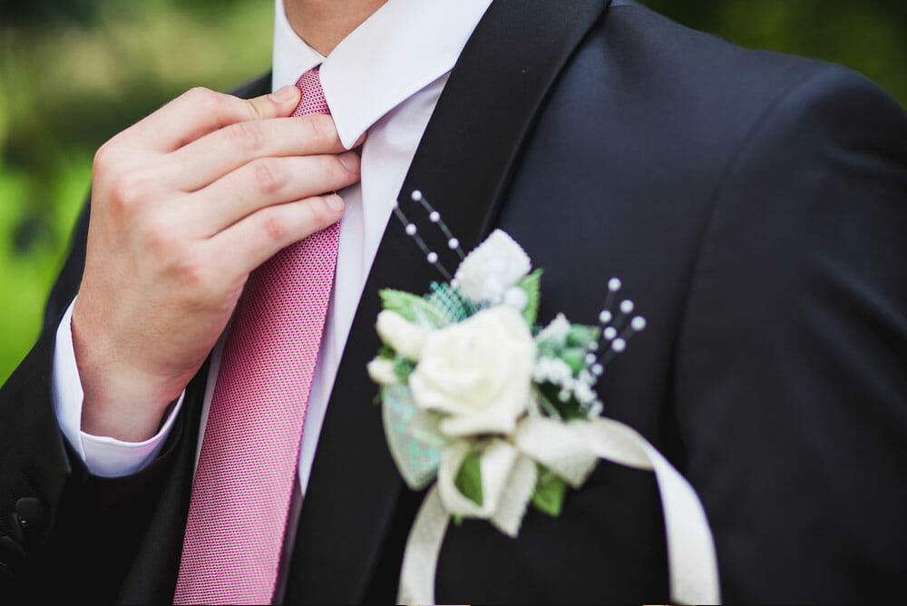 Close-up of a young man adjusting his pink tie while wearing a black suit and white dress shirt, with a white rose boutonniere pinned to his lapel for a formal event.