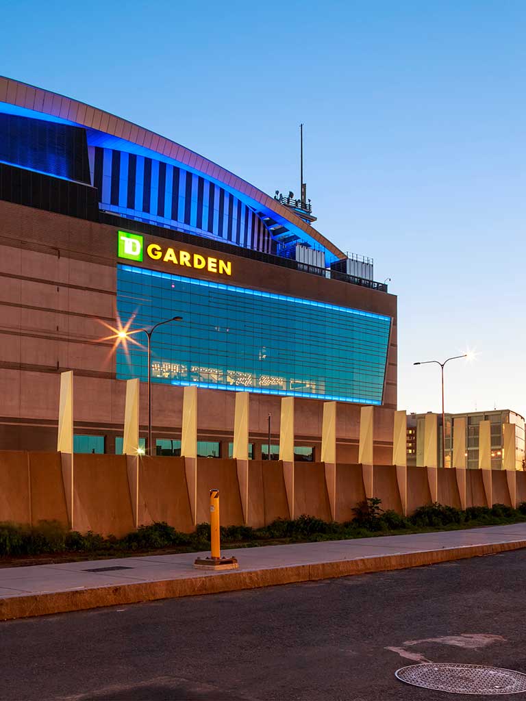 Exterior view of TD Garden in Boston at sunset with the Zakim Bridge in the background, both landmarks illuminated with lights.